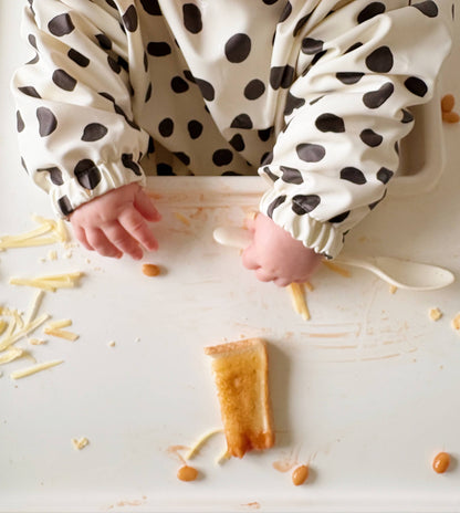 Child's hands reaching for food on a white surface with a polka dot bib.