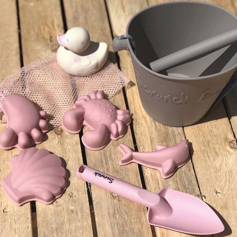 Pink sand toys including a shovel, molds, and a bucket on a wooden surface.