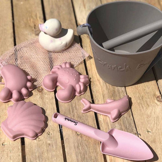 Pink sand toys including a shovel, molds, and a bucket on a wooden surface.