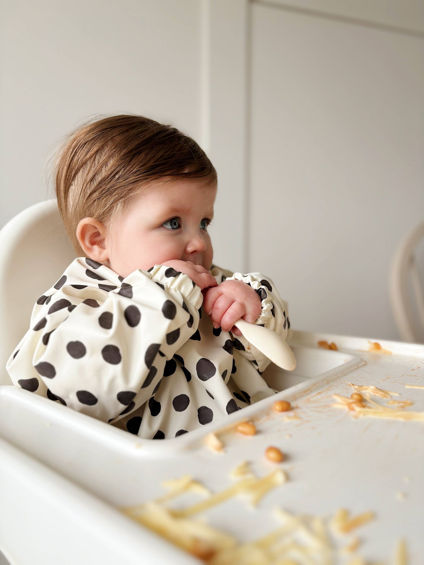 Baby in a high chair with a polka dot bib, surrounded by food on a white background