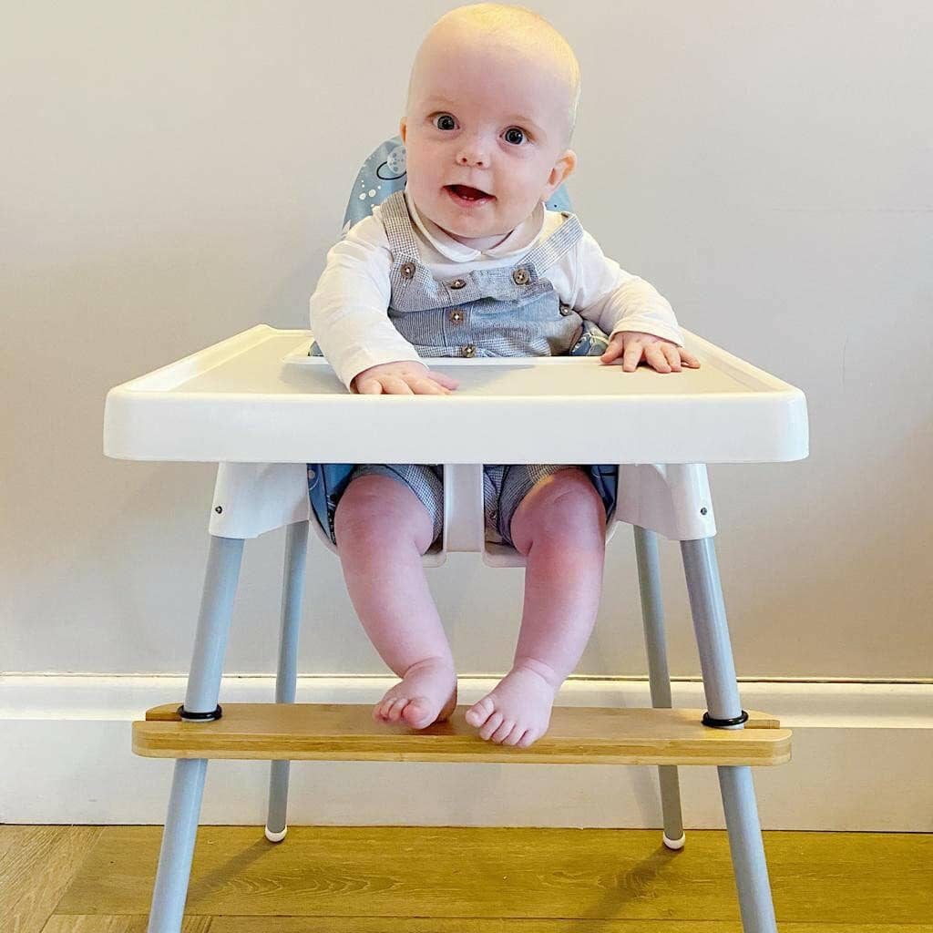 Baby sitting on a high chair with a wooden footrest attachment against a plain wall.