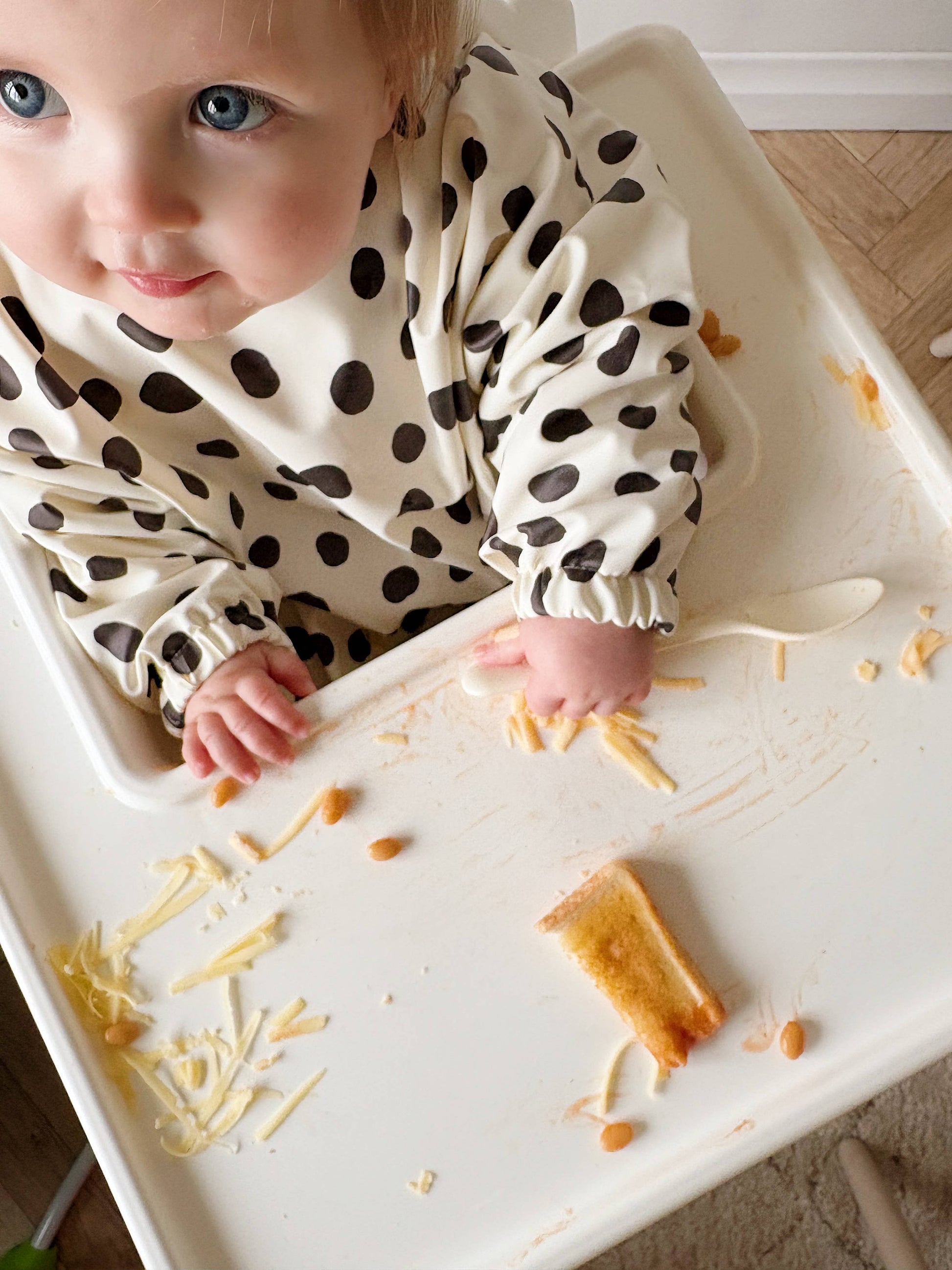 Baby in a polka dot bib sitting in a high chair with food on the tray