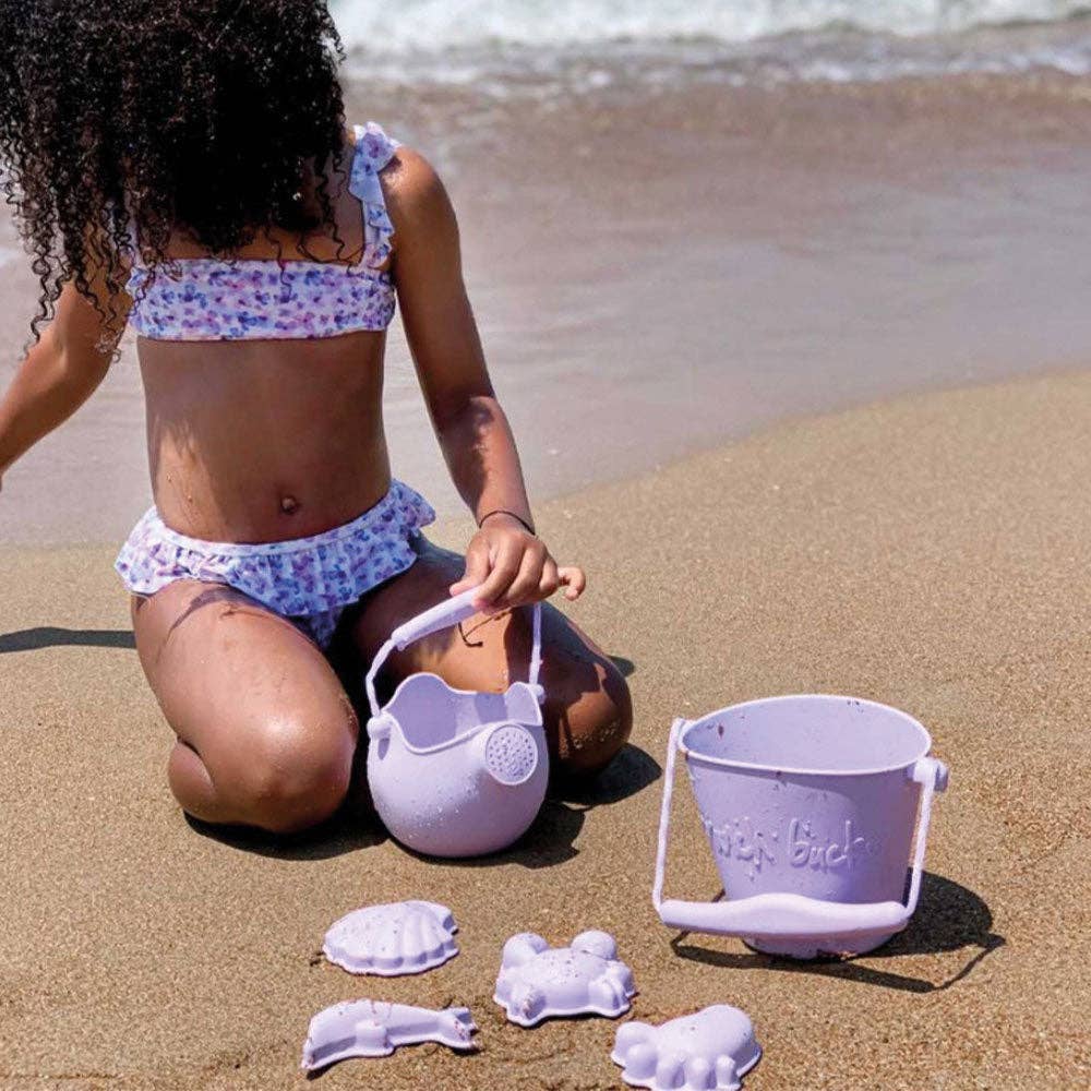 Child playing on a beach with sand toys, including a purple bucket and molds.