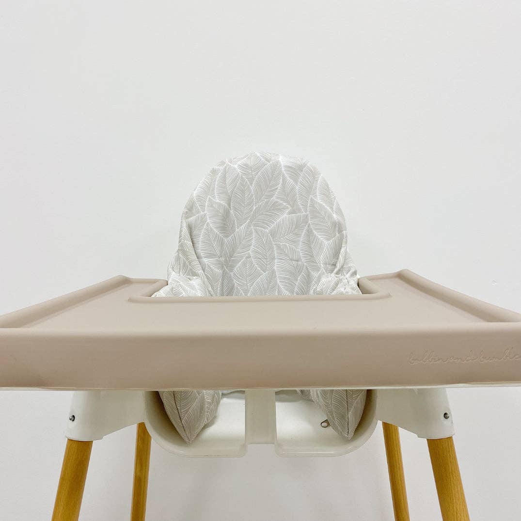 Beige silicone high chair tray cover with a white leaf patterned cushion on a plain background