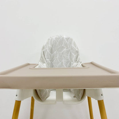 Beige silicone high chair tray cover with a white leaf patterned cushion on a plain background