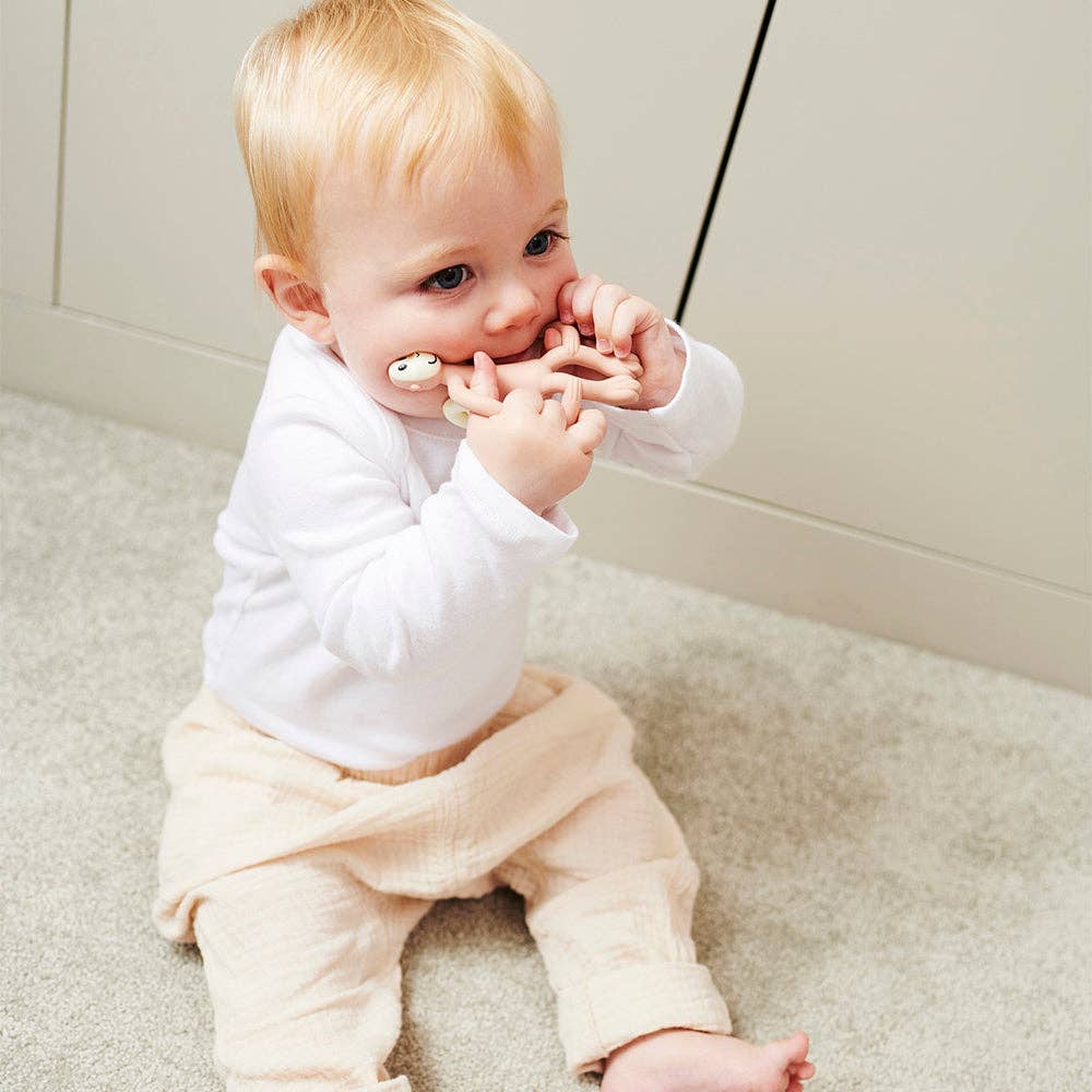 Baby sitting on a carpeted floor with a white shirt and beige pants, holding a white object.