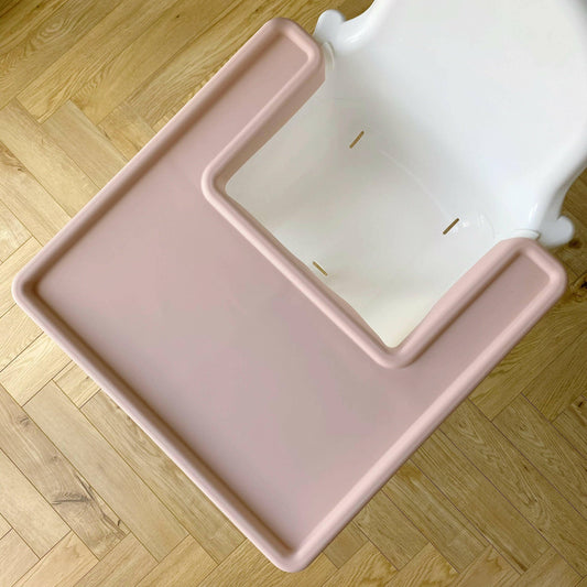 Pink silicone highchair tray cover on a white IKEA highchair, wooden floor in background